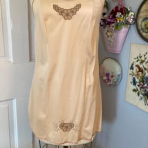 May include: A peach-colored slip with thin straps and a delicate floral embroidery. The slip is displayed on a mannequin, showcasing its vintage design and soft, flowing fabric. The trim is a darker peach color.