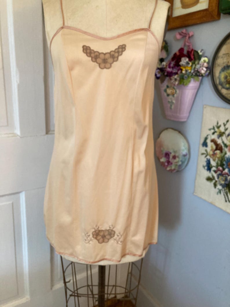 May include: A peach-colored slip with thin straps and a delicate floral embroidery. The slip is displayed on a mannequin, showcasing its vintage design and soft, flowing fabric. The trim is a darker peach color.