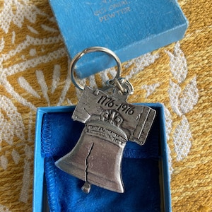May include: A silver-colored Liberty Bell keychain with the date 1776-1976 engraved on it, resting in a blue box. The box is labeled "Colonial Pewter". The keychain is on a yellow patterned surface.