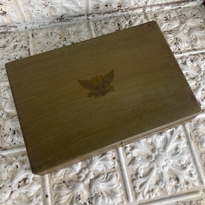 May include: A rectangular, olive-green wooden box with a gold emblem of an eagle on the lid. The box is closed and sits on a white, textured surface with a floral pattern. The box appears to be vintage.