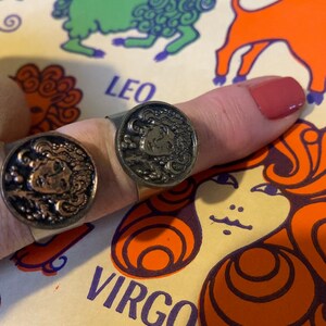 May include: Two gold-toned rings featuring zodiac symbols. The rings depict a woman's face with intricate hair and a crown. The rings are worn on a finger against a backdrop of a zodiac chart with the words "Leo" and "Virgo".