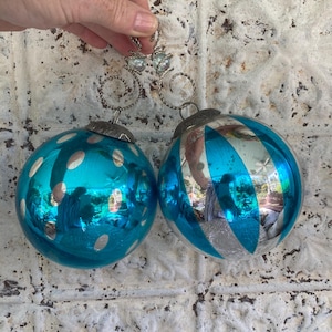May include: Two turquoise glass Christmas ornaments. One has silver polka dots, the other has silver stripes. Both have silver metal caps and hangers. The ornaments are round and reflective, with a vintage aesthetic.