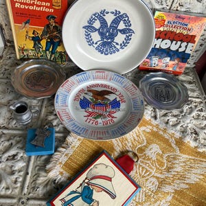 May include: A collection of vintage items including plates, books, and decorative pieces. The plates feature patriotic and commemorative designs. The books are titled "The American Revolution" and "Election Collection School House". A small metal pitcher and a framed illustration are also present.