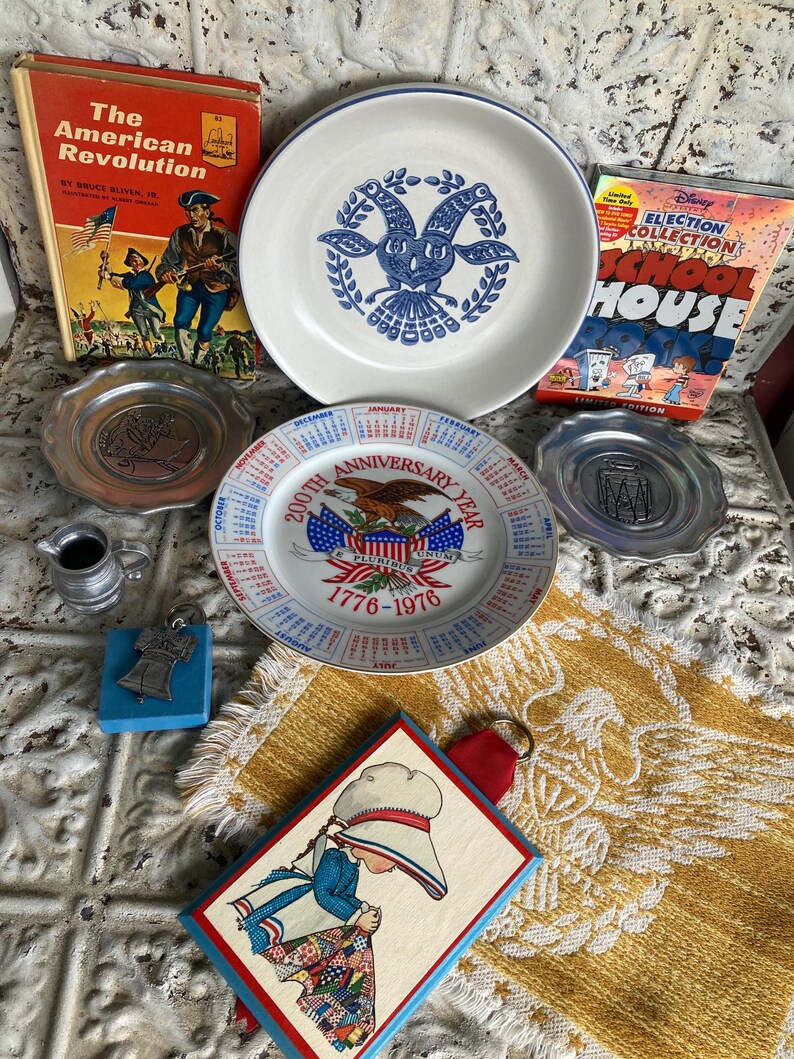 May include: A collection of vintage items including plates, books, and decorative pieces. The plates feature patriotic and commemorative designs. The books are titled "The American Revolution" and "Election Collection School House". A small metal pitcher and a framed illustration are also present.