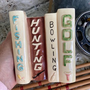 May include: A cream-coloured ceramic book-shaped vase featuring the words "FISHING", "HUNTING", "BOWLING", and "GOLF". The vase has brown and green accents, with illustrations of a fishing lure, a rifle, a bowling pin, and a golf tee.