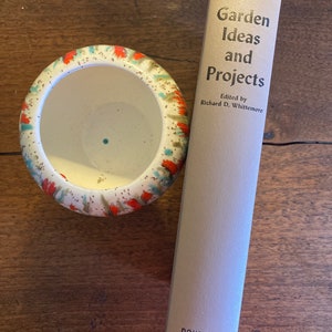 Puede incluir: Un libro vintage titulado "Garden Ideas and Projects" de Richard D. Whittemore, publicado por Doubleday. El libro está sobre una superficie de madera junto a una pequeña maceta de cerámica blanca con un diseño colorido.