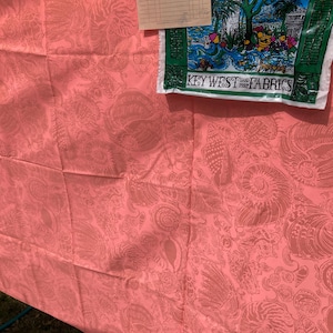 May include: Coral-colored fabric with a seashell pattern. A green and white bag with the text "Key West Fabrics" and a picture of a lighthouse is on top of the fabric. A paper receipt is also on top of the fabric.