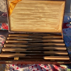 May include: An open antique cutlery case with a set of knives. The knives have light-colored handles and silver blades. The case is lined with gold fabric and has a brown exterior. The case is open, revealing the knives neatly arranged inside.