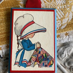 May include: A rectangular wooden plaque with a vintage illustration of a girl in a bonnet and blue dress, sewing a quilt. The plaque has a red border, blue edges, and a red ribbon for hanging.