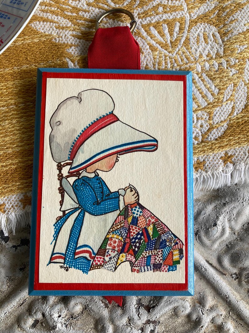 May include: A rectangular wooden plaque with a vintage illustration of a girl in a bonnet and blue dress, sewing a quilt. The plaque has a red border, blue edges, and a red ribbon for hanging.