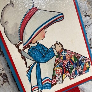 May include: A vintage wooden bookmark featuring a hand-painted illustration of a girl in a blue dress and white bonnet, holding a colorful patchwork quilt. The bookmark has a red ribbon and a blue and red border.