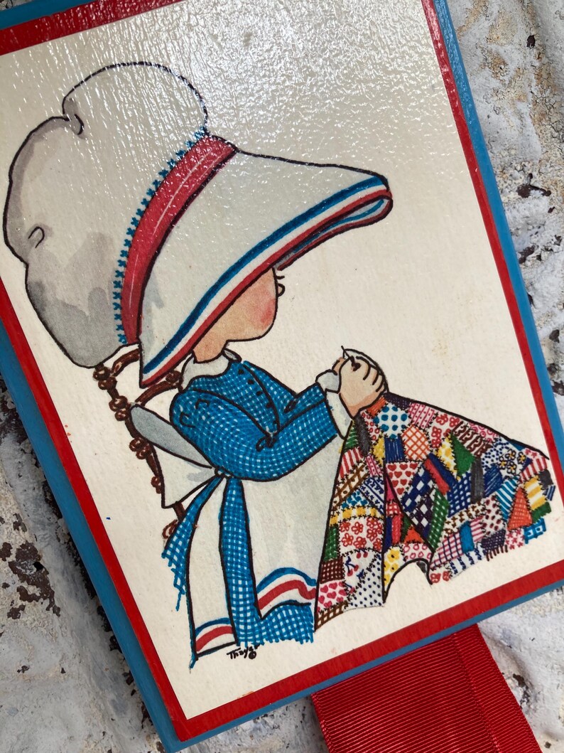 May include: A vintage wooden bookmark featuring a hand-painted illustration of a girl in a blue dress and white bonnet, holding a colorful patchwork quilt. The bookmark has a red ribbon and a blue and red border.