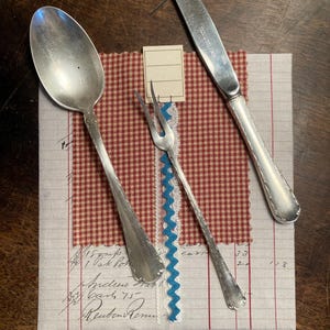 Puede incluir: Una cuchara plateada, un tenedor de dos puntas y un cuchillo de acero inoxidable dispuestos sobre una tela con patrón de cuadros rojos y blancos, un borde de cinta zigzag azul y una etiqueta de papel rectangular con tres líneas.