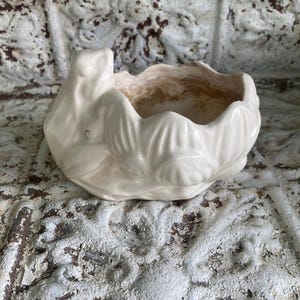 May include: A cream-colored ceramic planter shaped like a lotus flower with a frog perched on the side. The exterior features sculpted petal details, while the interior shows a light brown, weathered patina finish within the basin.