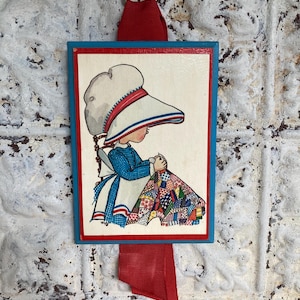 May include: A framed illustration of a child sewing a quilt, with a blue border and red ribbon. The child wears a blue dress, white apron, and a large white hat with red and blue stripes. The quilt is multi-colored.
