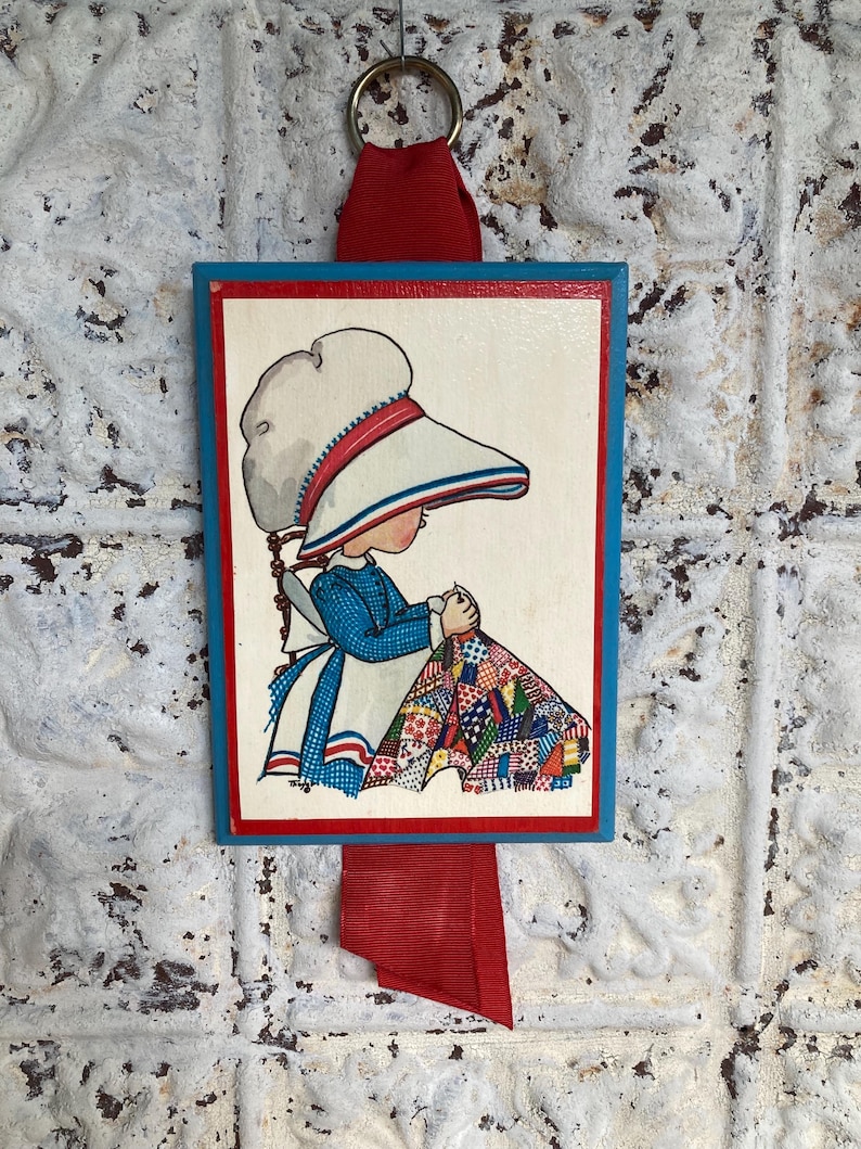 May include: A framed illustration of a child sewing a quilt, with a blue border and red ribbon. The child wears a blue dress, white apron, and a large white hat with red and blue stripes. The quilt is multi-colored.