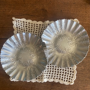 May include: Two silver-coloured plates with a ruffled edge and embossed floral patterns. They sit on a cream-coloured, crocheted doily, on a wooden surface. The plates have a decorative, vintage aesthetic.