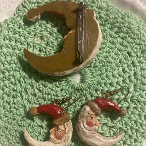 May include: A set of Christmas earrings and a brooch featuring a crescent moon with a Santa Claus face. The earrings and brooch are made of painted wood and have a rustic, vintage look.