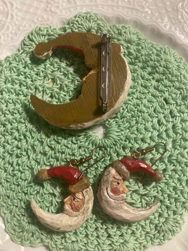 May include: A set of Christmas earrings and a brooch featuring a crescent moon with a Santa Claus face. The earrings and brooch are made of painted wood and have a rustic, vintage look.