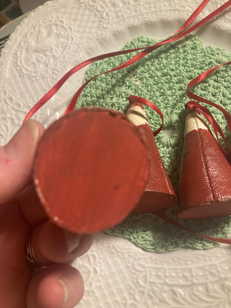 May include: Two red fabric cone-shaped ornaments with white trim and red ribbon ties. The ornaments are on a green crocheted surface.