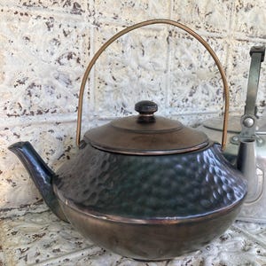 May include: Antique dark green and bronze tea kettle with a hammered texture. The kettle has a bronze handle and lid, and a black knob. The spout is dark green. A vintage kitchenware item.