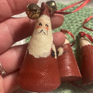 May include: Three red felt Christmas ornaments with painted Santa faces and gold bells. The ornaments are hanging from red ribbon.
