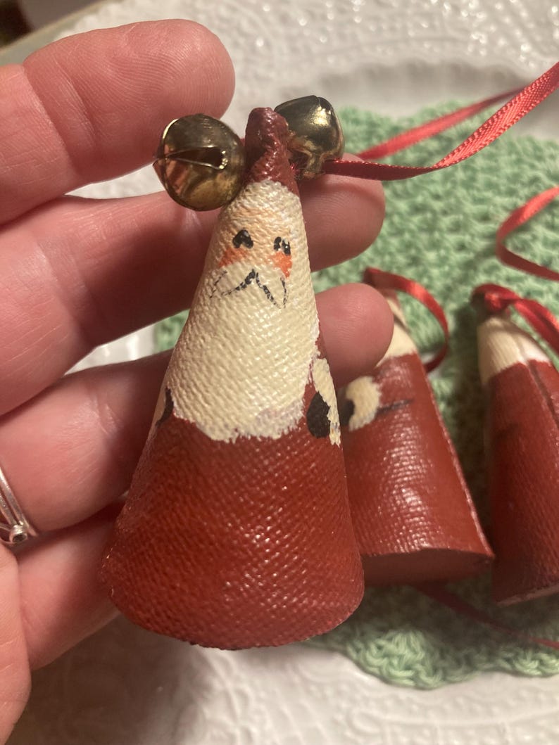 May include: Three red felt Christmas ornaments with painted Santa faces and gold bells. The ornaments are hanging from red ribbon.
