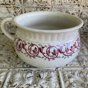 May include: A cream-colored ceramic chamber pot featuring a rounded body, a single side handle, and a decorative band of dark red floral and vine patterns encircling the exterior. The surface has a visible crackled glaze finish throughout.
