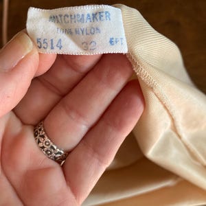 May include: A close-up of a tag from a garment, with the brand name "MATCHMAKER" printed in blue. The tag also indicates "100% NYLON", size "32", and "6FT". The garment is a light peach color.