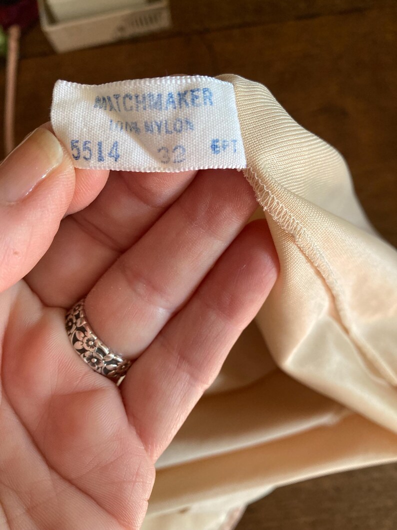 May include: A close-up of a tag from a garment, with the brand name "MATCHMAKER" printed in blue. The tag also indicates "100% NYLON", size "32", and "6FT". The garment is a light peach color.