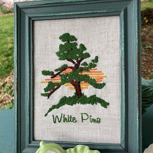 Può includere: Un ricamo incorniciato di un pino verde con un tramonto arancione alle sue spalle. Il ricamo è su uno sfondo di lino bianco e la cornice è di un verde blu scuro. Il testo "White Pine" è ricamato sotto l'albero.