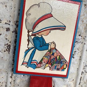 May include: A rectangular wooden plaque featuring a vintage illustration of a girl sewing a quilt. The artwork has a white background with a red and blue border. A red ribbon is attached to the top.