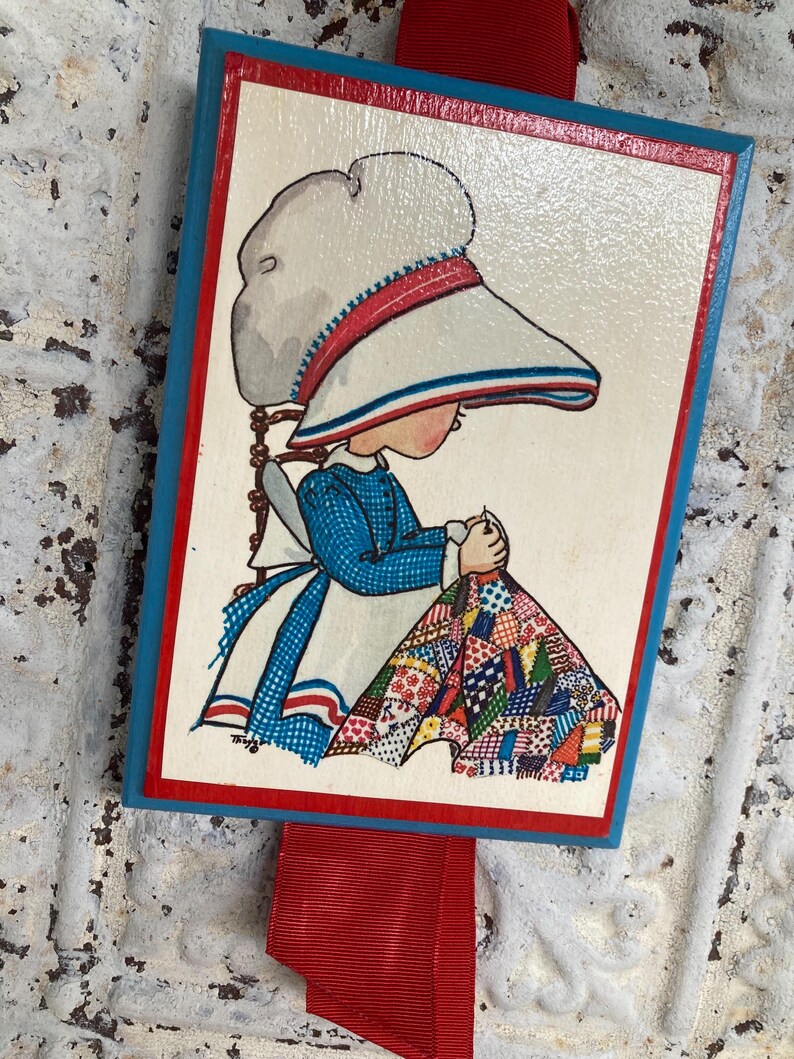 May include: A rectangular wooden plaque featuring a vintage illustration of a girl sewing a quilt. The artwork has a white background with a red and blue border. A red ribbon is attached to the top.