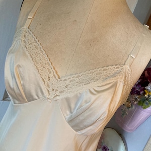 Op de afbeelding: Ivoor satijnen slip met delicate kanten afwerking langs de halslijn. De slip heeft verstelbare bandjes en een klassiek, elegant ontwerp. Het kledingstuk wordt op een mannequin getoond.