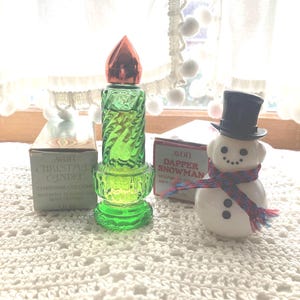 May include: Three vintage Avon cologne bottles. A green glass bottle shaped like a Christmas candle with a red top. A white ceramic bottle shaped like a snowman with a black top hat and red and blue scarf. A box for the Christmas Candle cologne with the text "Avon Christmas Candle Charisma Cologne Inspired by the fragrance of a white Christmas 1 fl oz (30 ml)". A box for the Dapper Snowman cologne with the text "Avon Dapper Snowman Moonwind Cologne 1 fl oz (30 ml)".