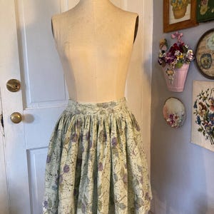 May include: A vintage-style, knee-length skirt displayed on a mannequin. The skirt features a light green background with a repeating pattern of purple floral and leaf designs. The fabric is gathered at the waist, creating a full, flared silhouette.