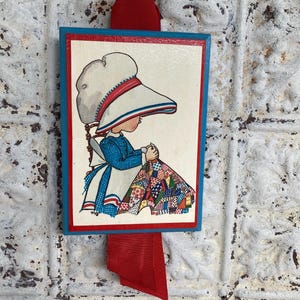 May include: A vintage wooden wall hanging featuring a framed illustration of a girl in a bonnet sewing a quilt. The frame has a blue border with a red inner border. A red ribbon and metal ring are attached for hanging.