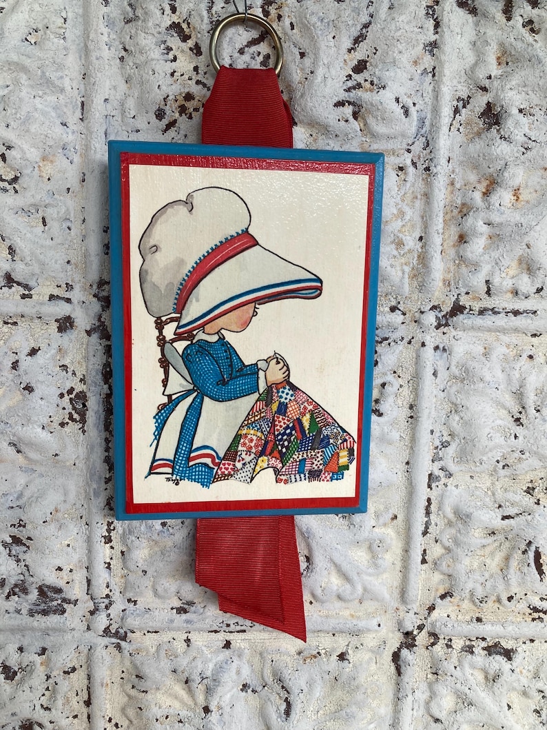May include: A vintage wooden wall hanging featuring a framed illustration of a girl in a bonnet sewing a quilt. The frame has a blue border with a red inner border. A red ribbon and metal ring are attached for hanging.