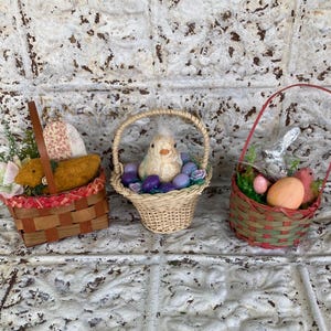 May include: Three decorative Easter baskets. The first basket is brown and red with a handle, containing a yellow item, flowers, and a patterned fabric. The second basket is woven, holding a chick and purple eggs. The third basket is red and green, with a silver bunny, eggs, and a pink bird.