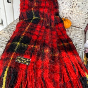 May include: A red, black, and yellow plaid scarf with fringe. The scarf has a tag that reads "PHYSICIAN Aero Rug". The scarf is draped over a white surface.