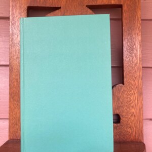 May include: A teal blue hardcover book with a smooth, textured cover. The book is sitting on a wooden shelf.