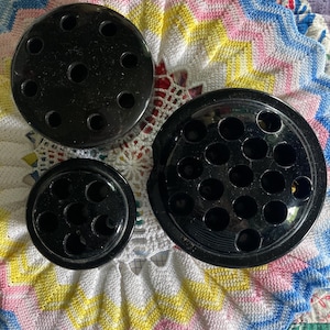 May include: Three black flower frogs of varying sizes. The frogs are round with multiple holes for arranging flowers. They are displayed on a colorful, patterned textile background.