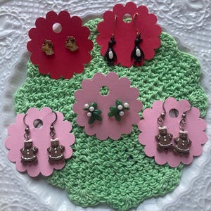 May include: Four pairs of Christmas-themed earrings on pink and red display cards. The earrings feature a gingerbread person, a penguin, a holly sprig with white pearls, and a gold bell.