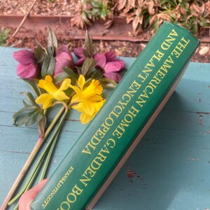 May include: A green hardcover book titled "The American Home Garden Encyclopedia and Plant Encyclopedia" by Evans &  Pringle. The book is on a blue surface with a bouquet of yellow and purple flowers.
