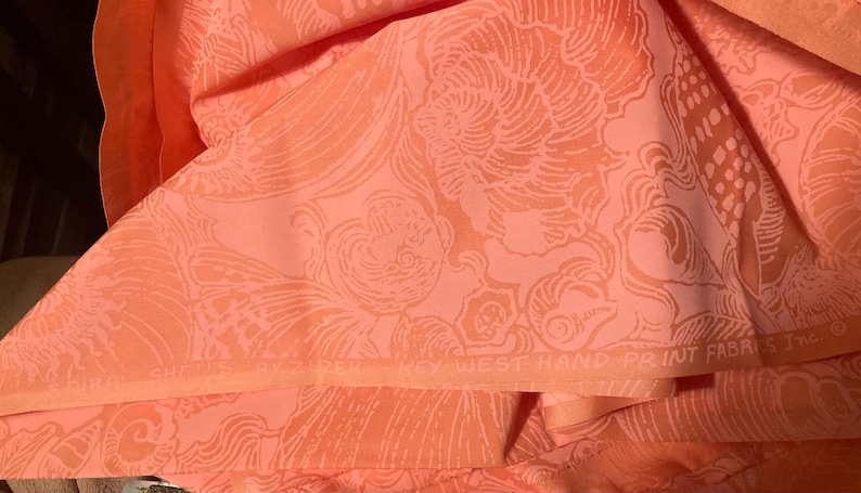 May include: Coral-colored fabric with a repeating pattern of stylized seashells and swirling lines. The fabric is folded, revealing the intricate design. The text "KEY WEST HAND PRINT FABRICS Inc" is visible.