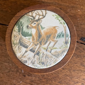 May include: A round wooden box with a ceramic top featuring a detailed illustration of a white-tailed deer in a forest setting. The deer is brown with white markings and has large antlers. The background is a green and brown forest scene.