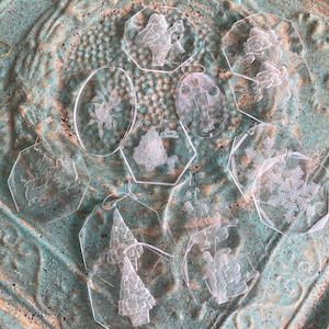 May include: A collection of clear, etched Christmas ornaments in various shapes. Designs include Santa Claus, Christmas trees, snowflakes, and floral patterns. Each ornament has a small hole for hanging. The ornaments are displayed on a textured, turquoise surface.