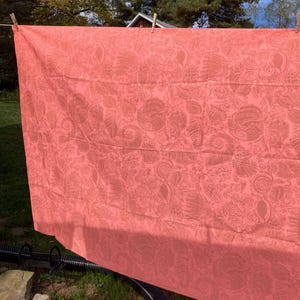 May include: A coral-colored textile with a detailed, repeating pattern of floral and abstract shapes. The fabric is hanging on a clothesline outdoors, with wooden clothespins securing it. The background includes green grass and trees.