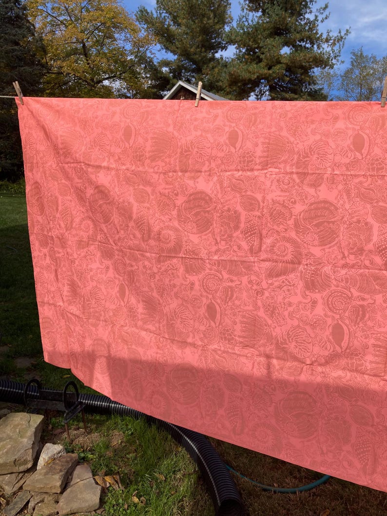 May include: A coral-colored textile with a detailed, repeating pattern of floral and abstract shapes. The fabric is hanging on a clothesline outdoors, with wooden clothespins securing it. The background includes green grass and trees.