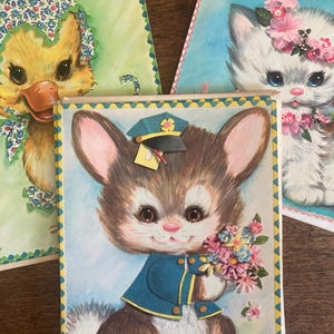 May include: Vintage greeting cards featuring adorable animal illustrations. The cards include a duckling in a bonnet, a kitten with a floral crown, and a bunny in a uniform holding flowers. The cards have decorative borders and are set against a wooden surface.